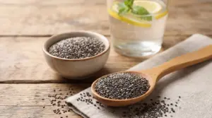 Semi di chia in cucchiaio e ciotola su tavolo di legno, accanto a un bicchiere d’acqua con limone
