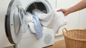 Mano che estrae asciugamani e camicia da un’asciugatrice aperta, accanto a un cesto della biancheria